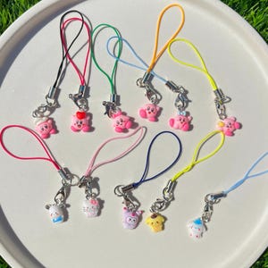 May include: A set of ten colourful phone charms featuring pink cartoon characters. The charms are attached to colourful strings and have silver clasps.