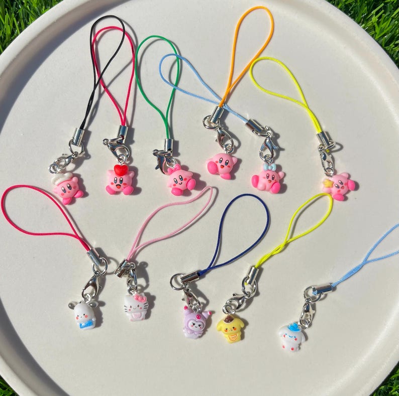 May include: A set of ten colourful phone charms featuring pink cartoon characters. The charms are attached to colourful strings and have silver clasps.