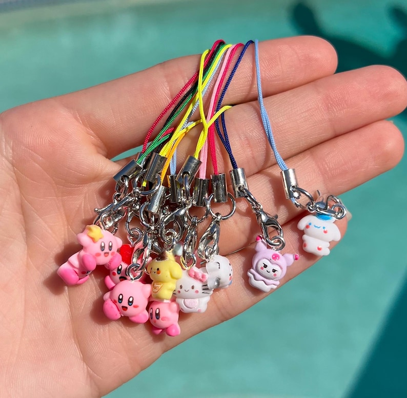 May include: A set of ten colorful phone charms featuring various cartoon characters. The charms are attached to colorful strings and have silver clasps. The charms include a pink and white character with a crown, a yellow character with a flower, a pink character with a star, a white character with a heart, a pink character with a bow, a white character with a flower, a pink character with a star, a white character with a heart, a pink character with a bow, and a white character with a flower.