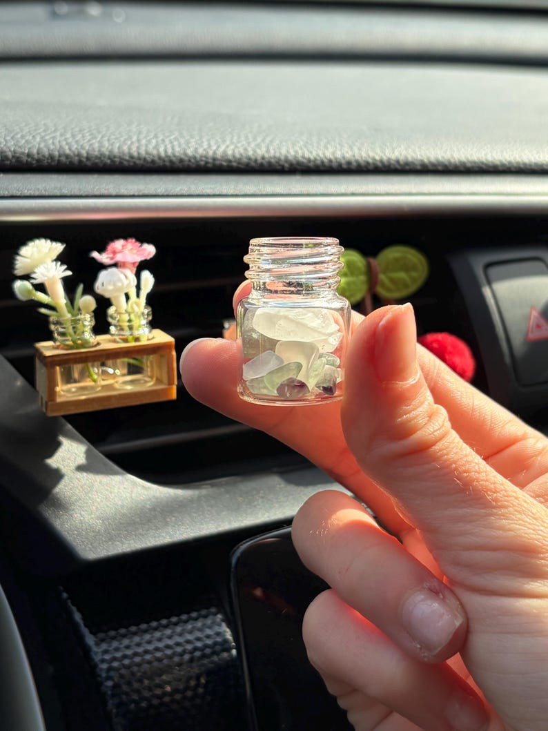 May include: A small, clear glass jar filled with sea glass and small purple stones. The jar is held in a hand, with a blurred background of a car's dashboard. Miniature floral arrangements are visible in the background.