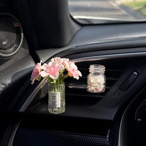 May include: A car interior with two small glass vases. One vase contains pink flowers, and the other contains pink petals. The vase with flowers has a label that reads "GOOD LUCK". The dashboard is black.
