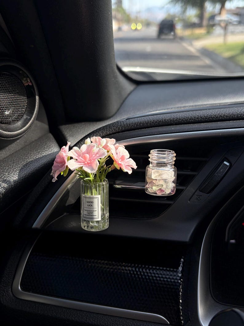 May include: A car interior with two small glass vases. One vase contains pink flowers, and the other contains pink petals. The vase with flowers has a label that reads "GOOD LUCK". The dashboard is black.