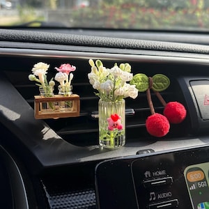 May include: Car air vent decorations featuring miniature flower arrangements in glass vases and a felt cherry ornament. The flowers include white and pink blooms. The cherry ornament has red felt cherries and green felt leaves.