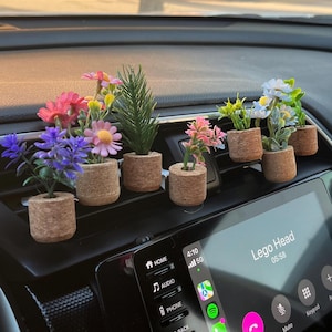 May include: A selection of miniature artificial flower arrangements in cork pots. The flowers are in shades of pink, purple, and white. The arrangements are placed on a car dashboard, providing a decorative element to the vehicle's interior.