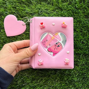 May include: A light pink mini binder featuring a heart-shaped cutout window displaying a graphic of a pink character with a bubble tea drink. The cover is decorated with small 3D character charms and includes a matching pink heart-shaped keychain.