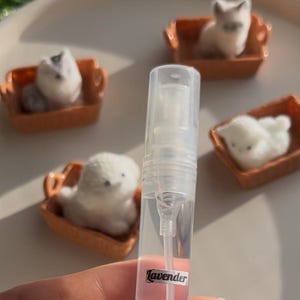 Puede incluir: Un frasco de spray de perfume transparente con una etiqueta "Lavender" se sostiene frente a figuras de gatos de fieltro en miniatura en sof&aacute;s en miniatura naranjas. Los gatos son blancos, grises y beiges. El fondo es de color beige claro.