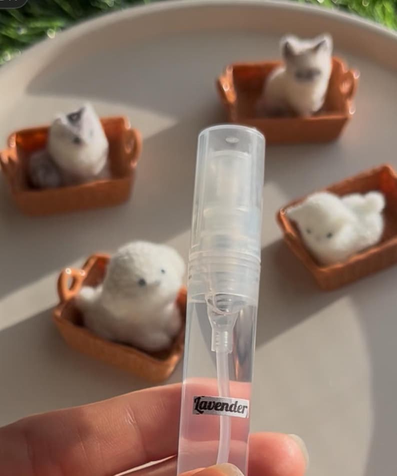 Puede incluir: Un frasco de spray de perfume transparente con una etiqueta "Lavender" se sostiene frente a figuras de gatos de fieltro en miniatura en sof&aacute;s en miniatura naranjas. Los gatos son blancos, grises y beiges. El fondo es de color beige claro.