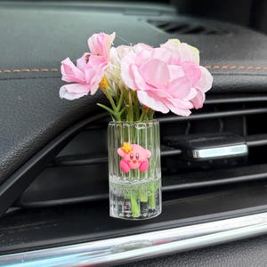 May include: A clear glass car vent clip with pink flowers and a cartoon character. The vase is filled with water and green stems. The pink flowers have multiple layers of petals.