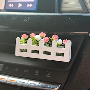 May include: A white picket fence-style car air vent decoration filled with pink and white artificial flowers. The decorative piece is placed in a car's dashboard, adding a touch of floral charm to the vehicle's interior.