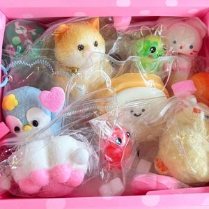 May include: A pink polka-dotted box filled with various colorful, soft, and squishy toys. The toys include a cat, an owl, a sushi roll, a green creature, and a white foot with pink toes, all individually packaged in clear plastic.