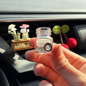 May include: A small, clear glass jar with a black and silver object inside, held in a hand. The jar is in focus, with a blurred background featuring a car's dashboard and decorative items, including a miniature wooden display with flowers.