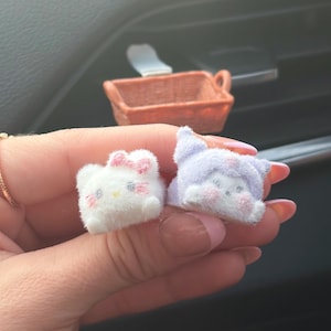 Puede incluir: Dos peque&ntilde;as figuras de personajes de peluche. Una es blanca con un lazo rosa, la otra es lavanda con una capucha. Ambas tienen rasgos faciales bordados. Una peque&ntilde;a cesta tejida naranja est&aacute; en el fondo.