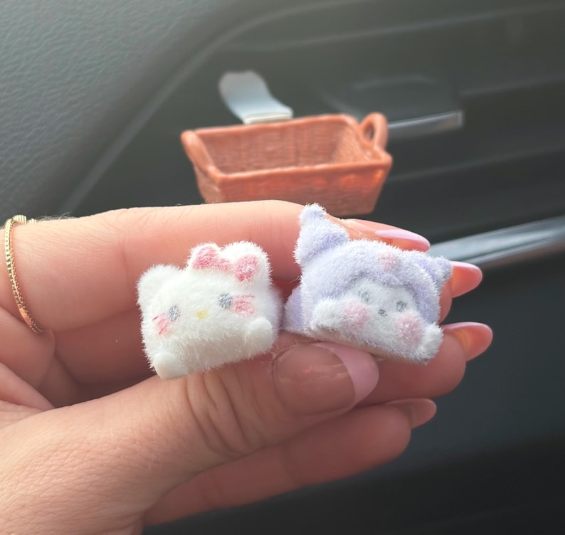 Puede incluir: Dos peque&ntilde;as figuras de personajes de peluche. Una es blanca con un lazo rosa, la otra es lavanda con una capucha. Ambas tienen rasgos faciales bordados. Una peque&ntilde;a cesta tejida naranja est&aacute; en el fondo.
