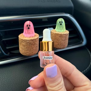May include: Two cylindrical cork car vent clips topped with small translucent ghost figures in pink and light green. A small clear glass dropper bottle with a gold collar and a white cap is held in the foreground, featuring a label that reads Jasmine.