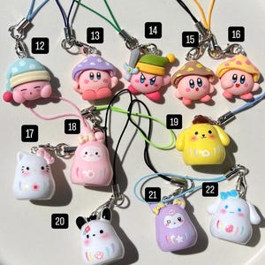 May include: A collection of 12 colourful, handmade phone charms featuring various characters, including a pink mushroom, a yellow dog, and a white cat. Each charm is attached to a string.