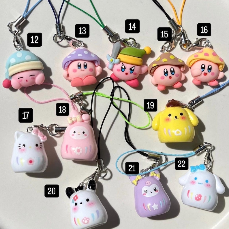 May include: A collection of 12 colourful, handmade phone charms featuring various characters, including a pink mushroom, a yellow dog, and a white cat. Each charm is attached to a string.