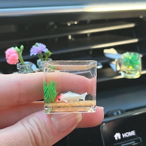 May include: A miniature, clear resin aquarium featuring a white and red koi fish, green grass, and sand. The small, square-shaped aquarium is held in a hand, with other miniature floral decorations in the background. The aquarium is a decorative item.