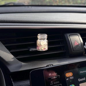 May include: A small, clear glass bottle filled with pink and white stones, placed on a car's air vent. The bottle has a silver-colored lid and is positioned in front of a black dashboard.