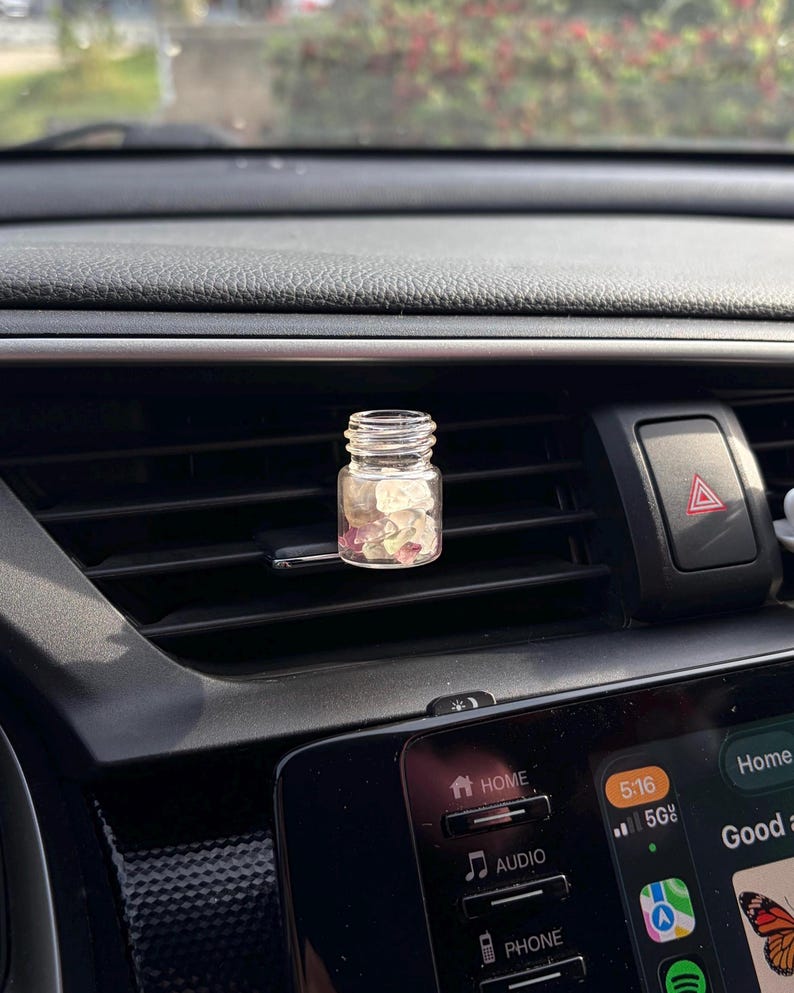 May include: A small, clear glass bottle filled with pink and white stones, placed on a car's air vent. The bottle has a silver-colored lid and is positioned in front of a black dashboard.
