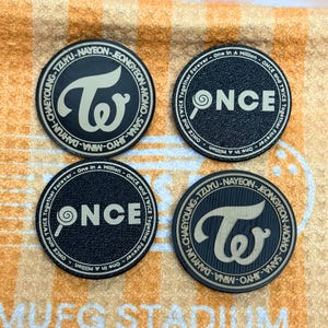 May include: Four black circular coasters with cream-colored text and logos. Two coasters feature the word "ONCE" with a lollipop graphic, and the other two display the band's logo and member names. The coasters rest on a textured orange and white surface.