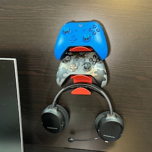 May include: A red wall mount with two video game controllers and a pair of black headphones. The controllers are a blue Xbox controller and a camouflage Xbox controller. The headphones have the text "SteelSeries" on the side.