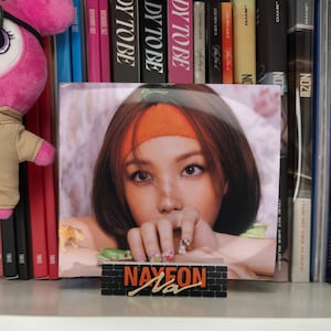 May include: A close-up of a CD featuring a woman with long brown hair wearing an orange headband. The CD is on a shelf with other CDs and books. The CD cover has the text "NAYEON" in large, bold letters.