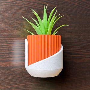 May include: A small, orange and white planter with a green succulent plant. The planter has a ribbed orange section and a white, speckled base and angled support. The plant has long, pointed green leaves.