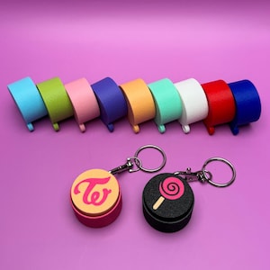 May include: A set of ten colorful round keychains with different designs. The keychains are arranged in a row on a pink surface. The keychains feature a pink and yellow keychain with the text "TWICE" and a black keychain with a pink lollipop design.