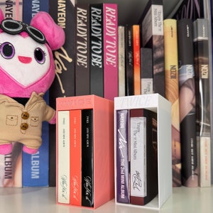 Peut inclure: Deux boîtes blanches et roses avec le mot "TWICE" imprimé dessus. Les boîtes sont remplies de CD et portent les titres d'album "Feel Special", "More & More", "Eyes Wide Open", "Formula of Love: O+T=<3", "The Story Begins", "TWICE", "The 2nd Mini Album", "The 1st Mini Album", et "TWICE" imprimés dessus.