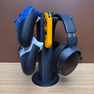 May include: Black gaming controller stand holding a blue and black controller, a white and black controller, a yellow controller, and a black headset. The stand is black and sits on a wooden surface.