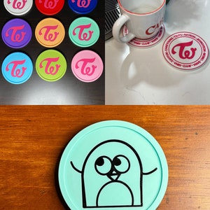 May include: A collection of colorful round coasters featuring a stylized logo in pink and white. A white mug with a red rim sits on a coaster with the names of the members of the group. A teal coaster with a black penguin illustration.