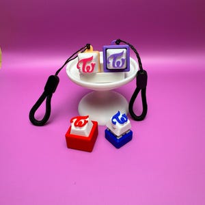 Puede incluir: Cuatro llaveros con el logotipo "TWICE" en diferentes colores: rosa, morado, rojo y azul. Los llaveros están en un soporte para pasteles blanco con un cordón negro.