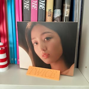 Könnte beinhalten: Nahaufnahme eines Twice-Albumcovers mit einer Frau mit dunklen Haaren und Make-up. Das Albumcover befindet sich auf einem Regal mit einem gelben Ständer, auf dem "TWICE" in fetten Buchstaben steht.