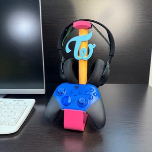 Puede incluir: Un soporte para auriculares de juego rosa y azul con un logotipo "TWICE" azul. El soporte sostiene un mando Xbox azul.