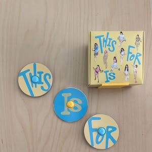 Puede incluir: Un juego de tres imanes redondos y una caja amarilla. Los imanes son amarillos con texto azul que dice "This", "Is" y "For". Cada imán tiene un botón de color en el centro. La caja tiene las palabras "This Is For" e ilustraciones de personas.