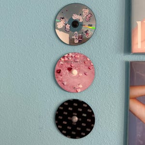 May include: Three CDs displayed on a light blue wall. The top CD has a silver background with cartoon animal stickers. The middle CD has a pink background with a pink and white heart pattern and the text "The Story of Love 01". The bottom CD has a black background with a white repeating pattern.