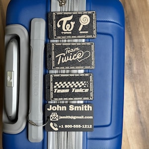 May include: Four rectangular black luggage tags with white text and graphics arranged vertically on a blue suitcase. The tags feature Twice branding, including the group name, member names, and a personalized tag with the name John Smith, email, and phone number.