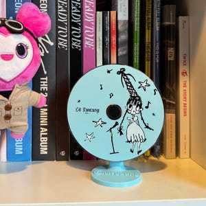 Może przedstawiać: Jasnoniebieski stojak w kształcie płyty CD z kreskówkową ilustracją dziewczyny, gwiazdami i nutami. Tekst "Lil Fantasy Vol. 1" jest wydrukowany na płycie CD. Stojak stoi na białej półce z książkami i pluszakiem.
