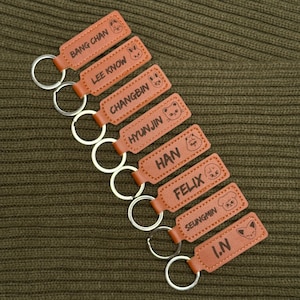 May include: A collection of brown leather keychains with silver key rings. Each keychain is rectangular and features a name and a small cartoon animal illustration. The names include "BANG CHAN", "LEE KNOW", "CHANGBIN", "HYUNJIN", "HAN", "FELIX", "SEUNGMIN", and "I.N".