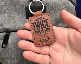 Op de afbeelding: Een bruine leren sleutelhanger met de tekst "TWICE THIS IS FOR WORLD TOUR 2026" in het zwart. De rechthoekige sleutelhanger is bevestigd aan een zilveren sleutelring. De achtergrond bevat een grijze rugzak.