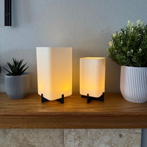 May include: Two white square pillar candles with black metal bases on a wooden shelf. The candles are lit and glowing.