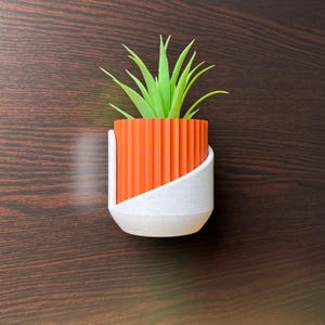 May include: A small, orange and white planter with a green succulent plant. The planter has a ribbed orange body and a white, angled support. The plant has long, pointed green leaves. The planter is set against a dark brown background.