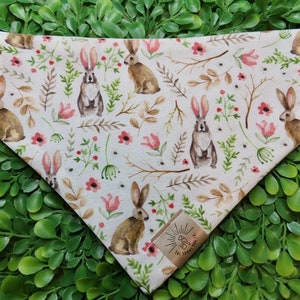 May include: A cream-colored dog bandana featuring a repeating pattern of watercolor-style rabbits, pink flowers, and green foliage. The bandana has a triangular shape and a small tag with text. The design evokes a spring or Easter theme.