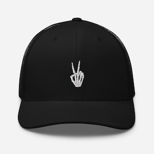 May include: Black trucker hat with a white skeleton hand making a peace sign. The hand has pink accents on the fingers.