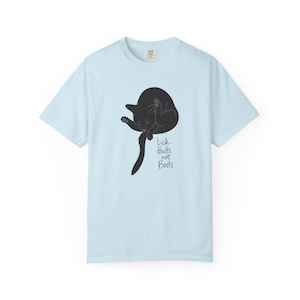 May include: Light blue t-shirt featuring a black cat grooming itself. The cat illustration is accompanied by the text "Lick Butts not Boots" in a handwritten style font. The shirt has a crew neck and short sleeves.