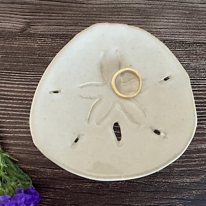 May include: A sand dollar-shaped ceramic ring dish in a light beige color, with a gold ring in the center. The dish has a starfish design and small holes. A purple flower and green stem are in the lower left corner.
