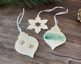 May include: Three white ceramic Christmas ornaments with white ribbons for hanging. One is a snowflake, and two are teardrop-shaped with cut-out designs and green accents. The ornaments are on a wooden surface.