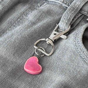 May include: A pink heart-shaped charm hangs from a silver-colored metal clasp. The clasp is attached to a gray denim fabric, likely a pocket or belt loop. The charm is a solid pink color.