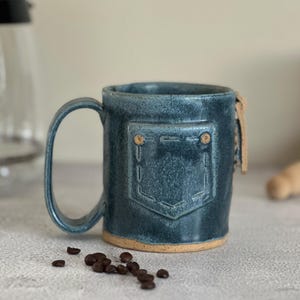May include: A blue ceramic mug with a textured finish featuring a molded pocket design on the front. The mug has a curved handle and a light brown base. The surface of the mug shows subtle variations in color and a stitched pattern detail.