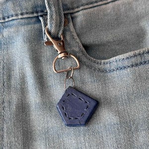 May include: A blue, diamond-shaped keychain with white stitching hangs from a silver carabiner clip attached to a light blue denim pocket. The keychain has a textured surface and a small metal ring.